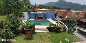 A swimming pool area with a blue building, steps leading to the pool, and a garden with a climbing frame. - Villa Pribadi Beunying 35 pax (Cipanas)