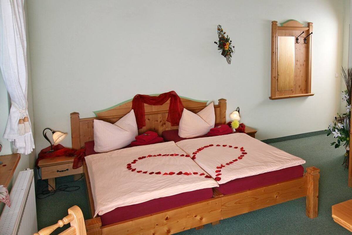 A wooden bed with rose petals arranged in heart shapes on the bedspread, a bedside lamp, and a mirror on the wall.