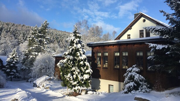 A wooden chalet with a sloped roof, surrounded by snow-covered trees and a mountain in the background. - Gasthof&Pension Brettmühle (Koenigswalde)