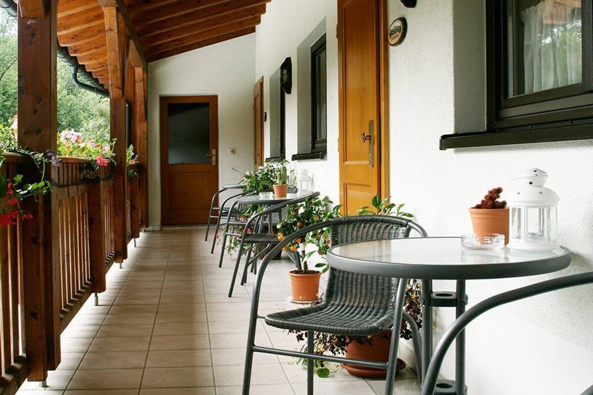 A covered outdoor area with a row of wicker chairs and tables, potted plants, and a wooden door.