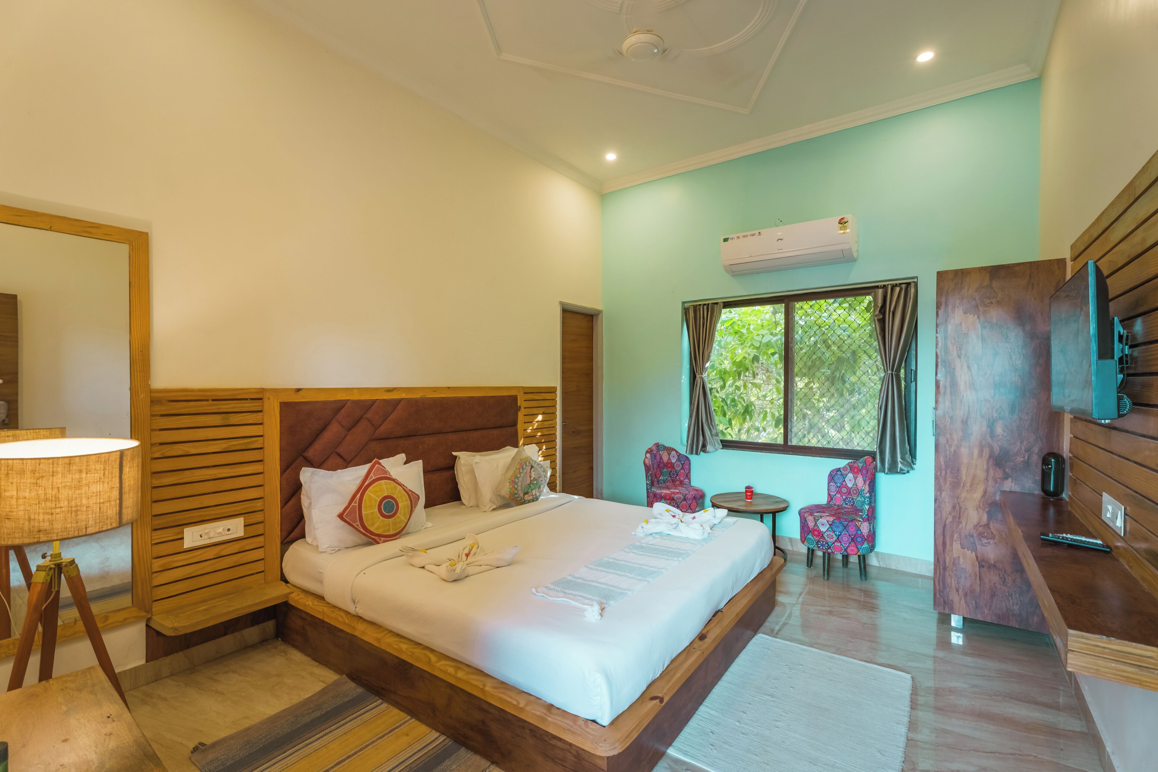 A hotel room with a large bed, a wooden headboard, a small table, two chairs, a TV, and an air conditioning unit.