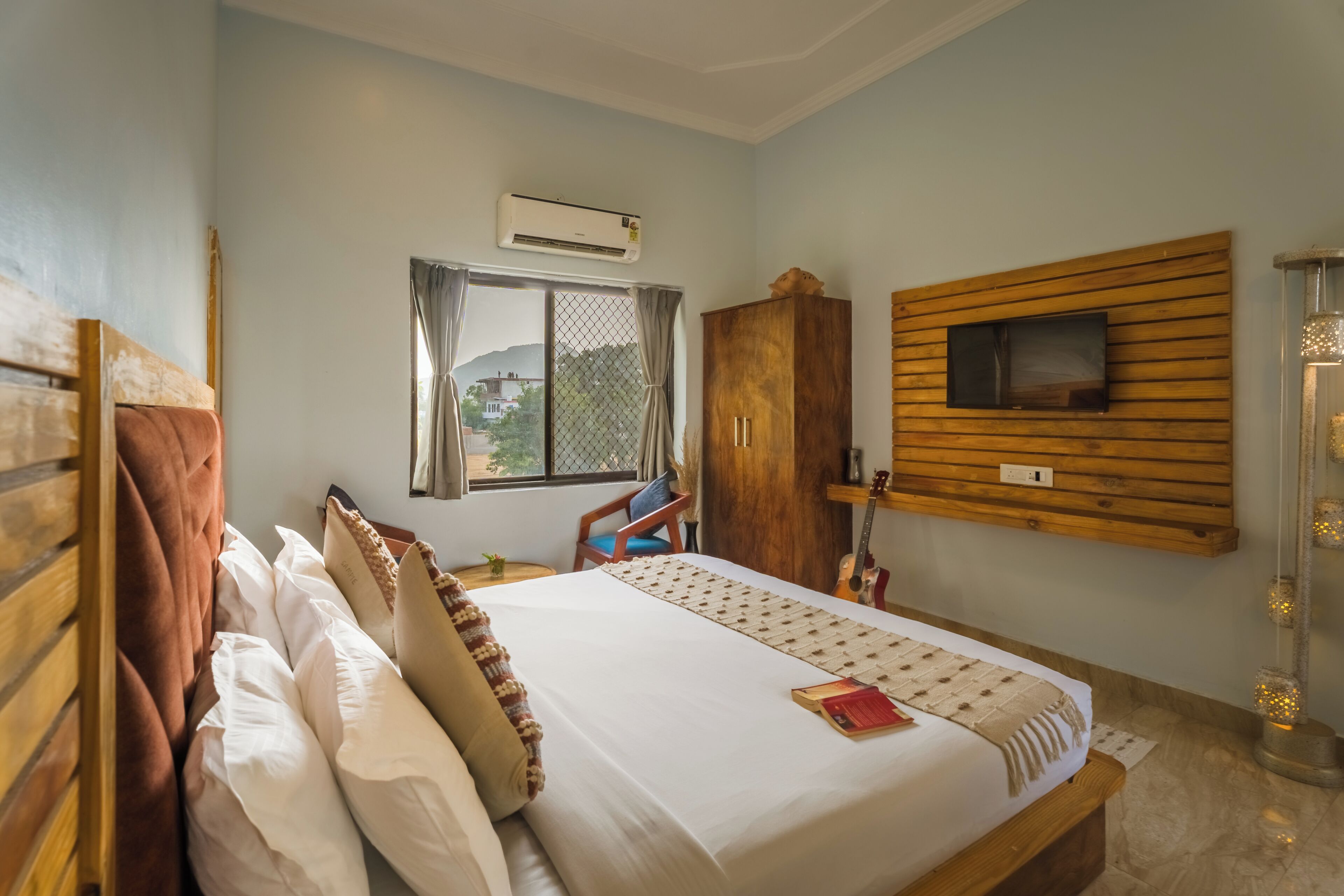 A hotel room with a bed, a TV mounted on the wall, a window with curtains, and a wooden wardrobe.