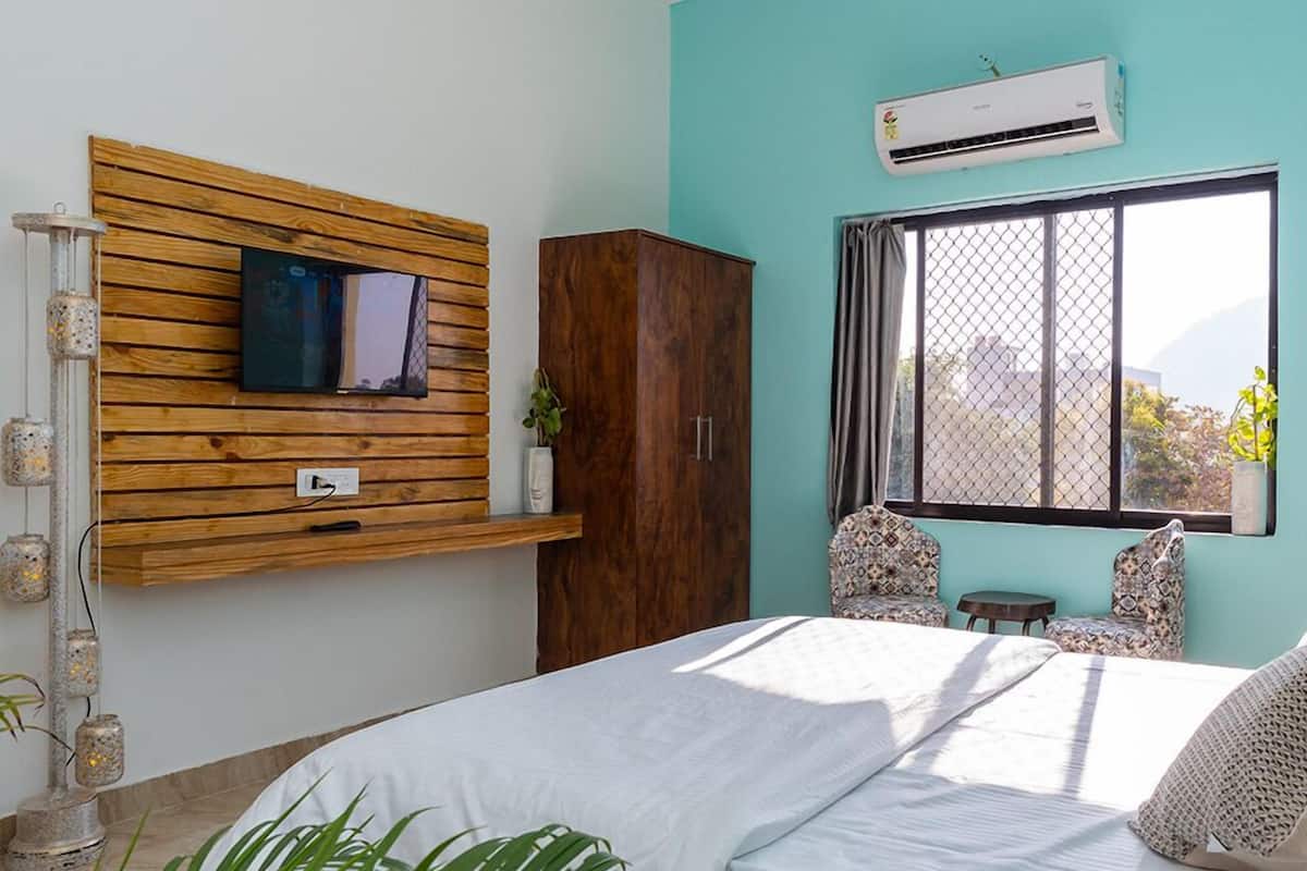 A bedroom with a bed, a TV mounted on a wooden panel, an air conditioning unit, and a window with a view.