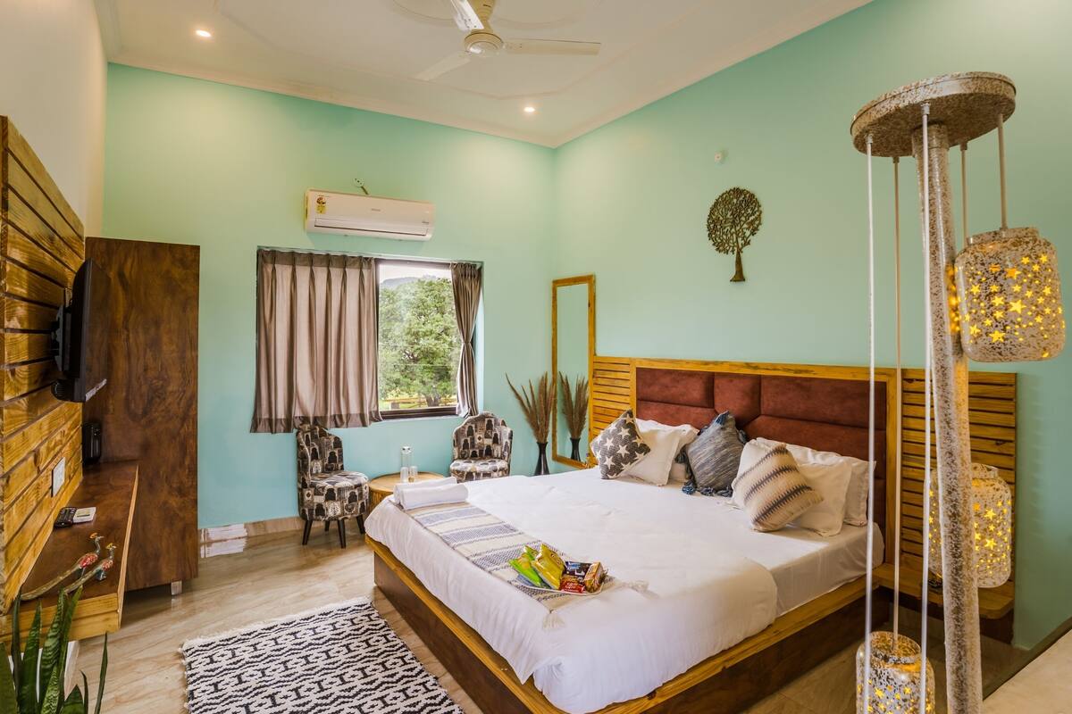 A bedroom with a large bed, a wooden headboard, a ceiling fan, an air conditioning unit, a window with curtains, a chair, and a decorative tree on the wall.