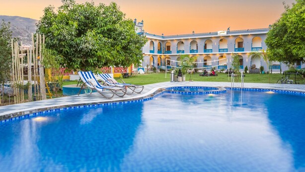 A swimming pool with lounge chairs, a large tree, and a building with arches in the background. - Moustache Pushkar - Hostel (Pushkar)