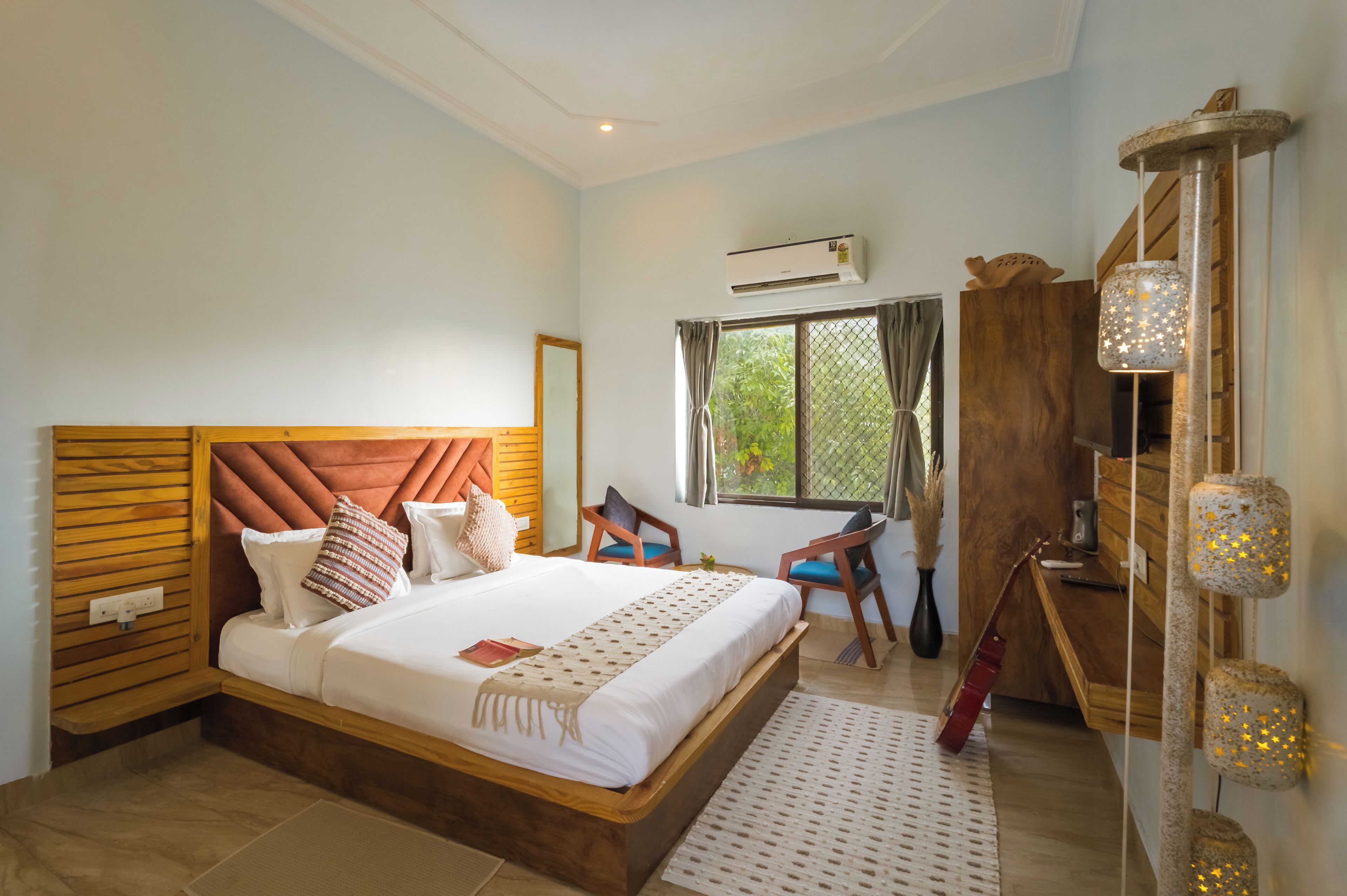 A bedroom with a large bed, a wooden headboard, a desk, a chair, a window with curtains, and a wall-mounted air conditioner.