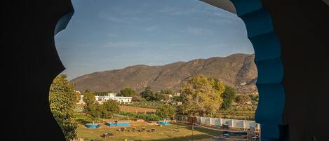 A view through an arched window of a backyard with a pool, outdoor furniture, and a mountainous landscape in the background.