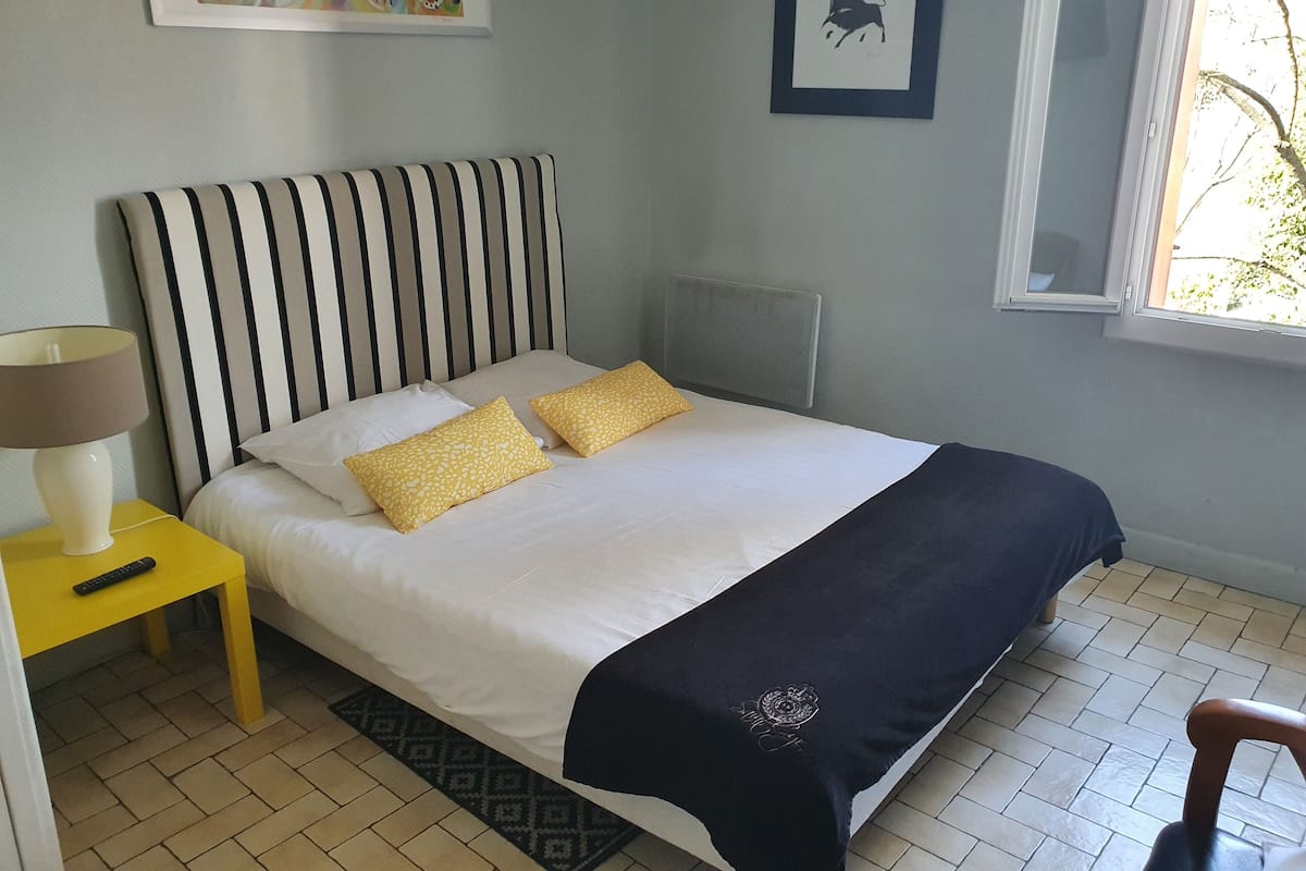 Uma cama bem arrumada com cabeceira listrada, duas almofadas, um cobertor preto e uma almofada amarela. Há uma mesa de cabeceira com uma luminária e um controle remoto. O quarto possui piso de azulejos e uma janela com vista para árvores.