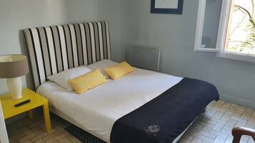A neatly made bed with a striped headboard, two pillows, a black throw blanket, and a yellow pillow. There is a bedside table with a lamp and a remote control. The room has a tiled floor and a window with a view of trees.