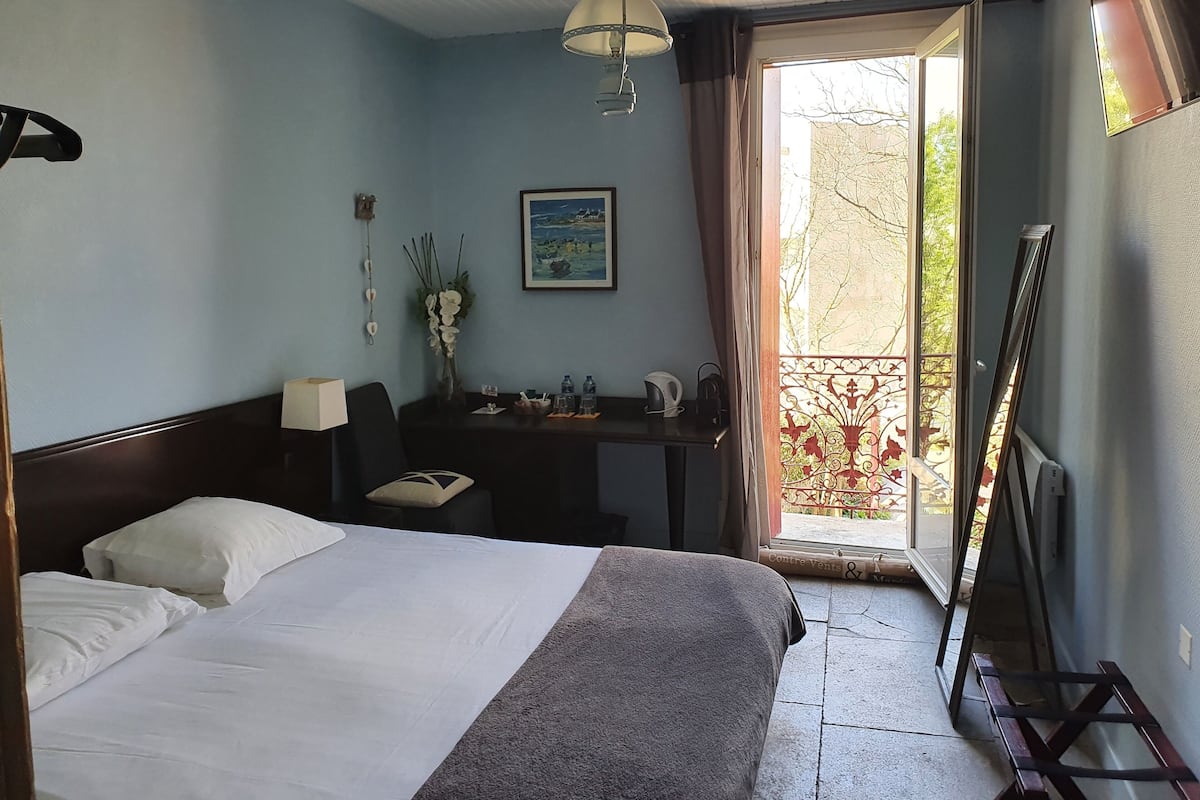 Quarto de hotel com uma cama, escrivaninha, cadeira, luminária, pintura e vista para o exterior.