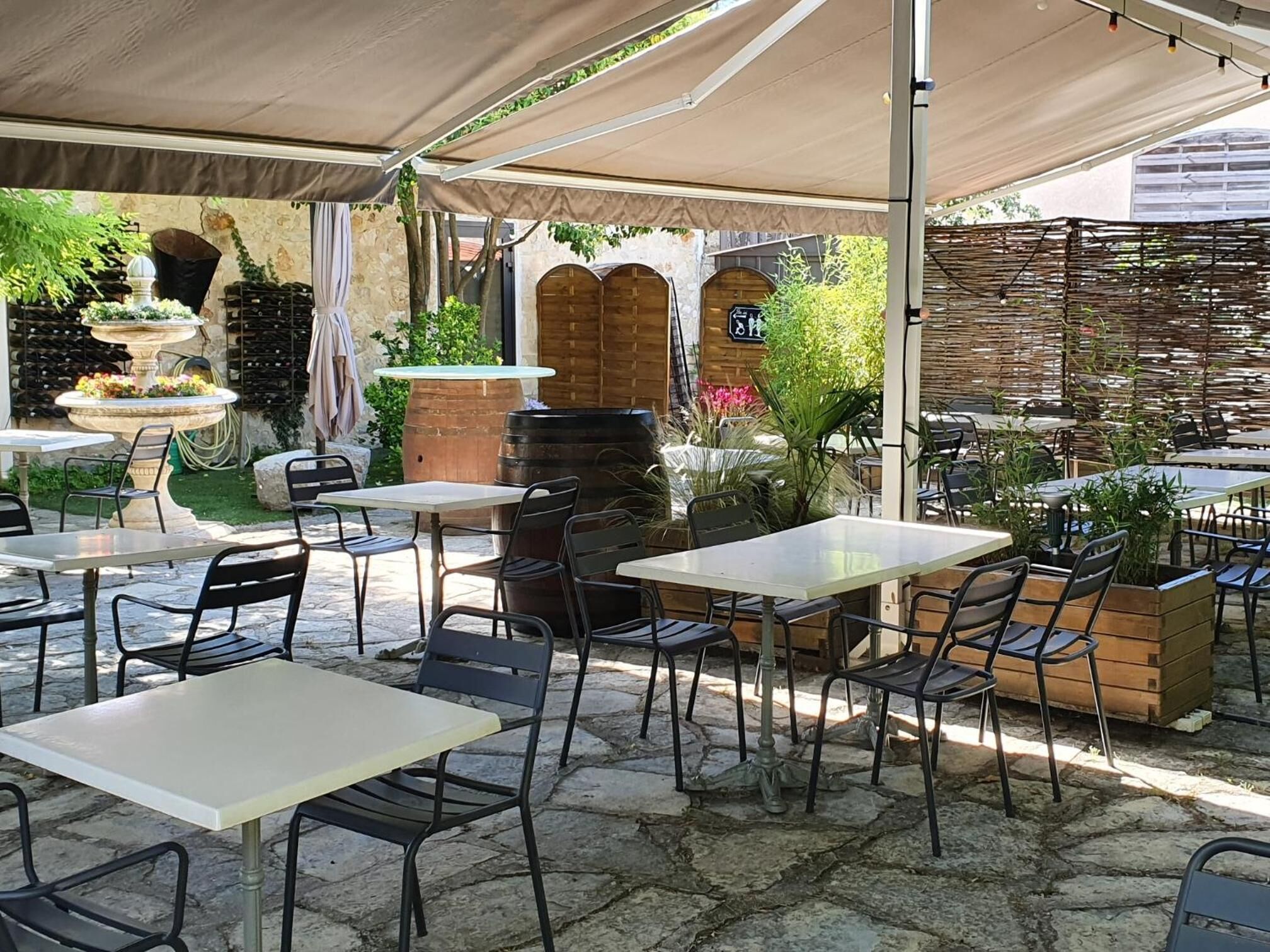 An outdoor patio with stone flooring, tables, and chairs. There are potted plants and a decorative fountain.