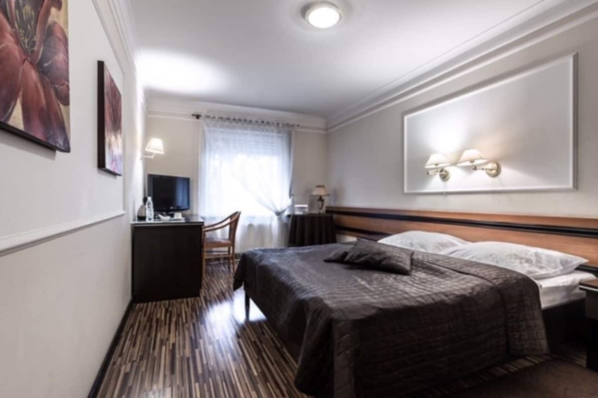 A hotel room with a bed, a desk, a chair, a television, and a wall-mounted light fixture.
