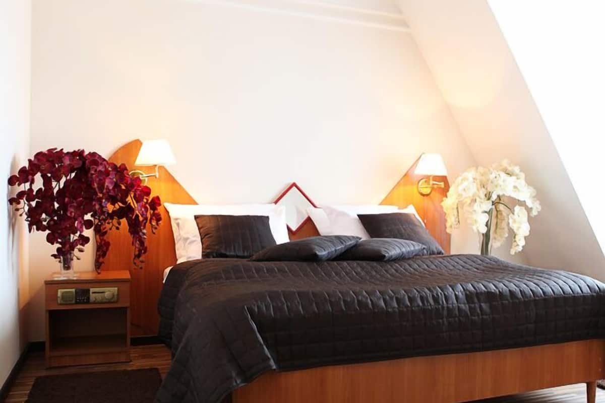 A bedroom with a sloped ceiling, a bed with a dark quilt, two bedside tables with lamps, and a wooden floor.