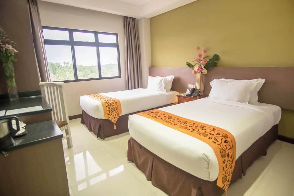 A hotel room with two beds, a desk with a telephone, and a window with a view of greenery.