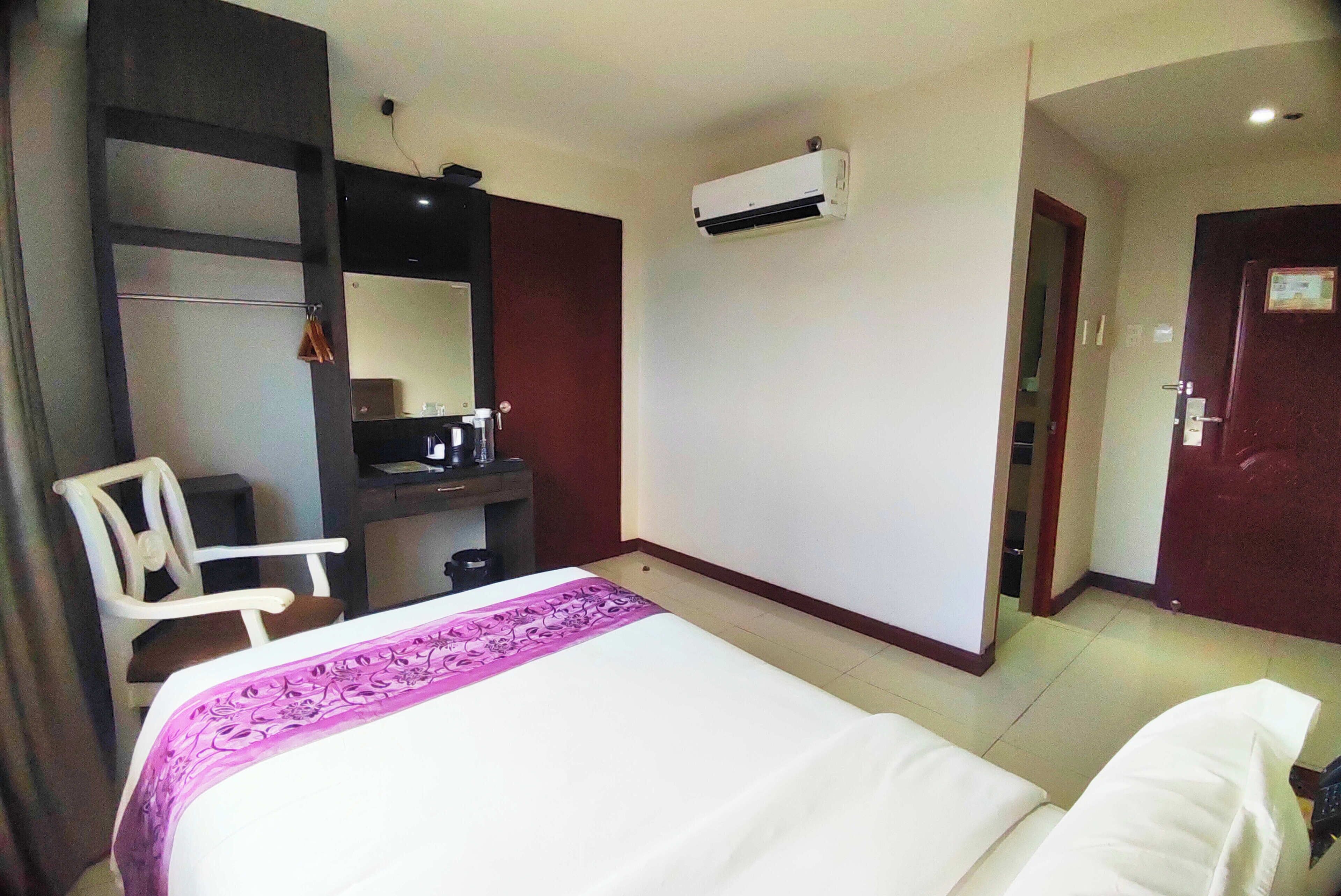 A hotel room with a bed, a desk, a chair, an air conditioning unit, and a door.