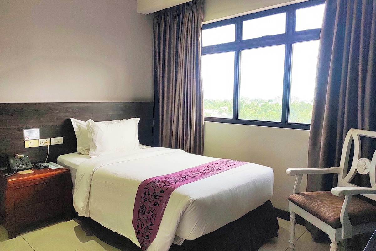A hotel room with a bed, a nightstand with a phone, a chair, and a window with curtains.