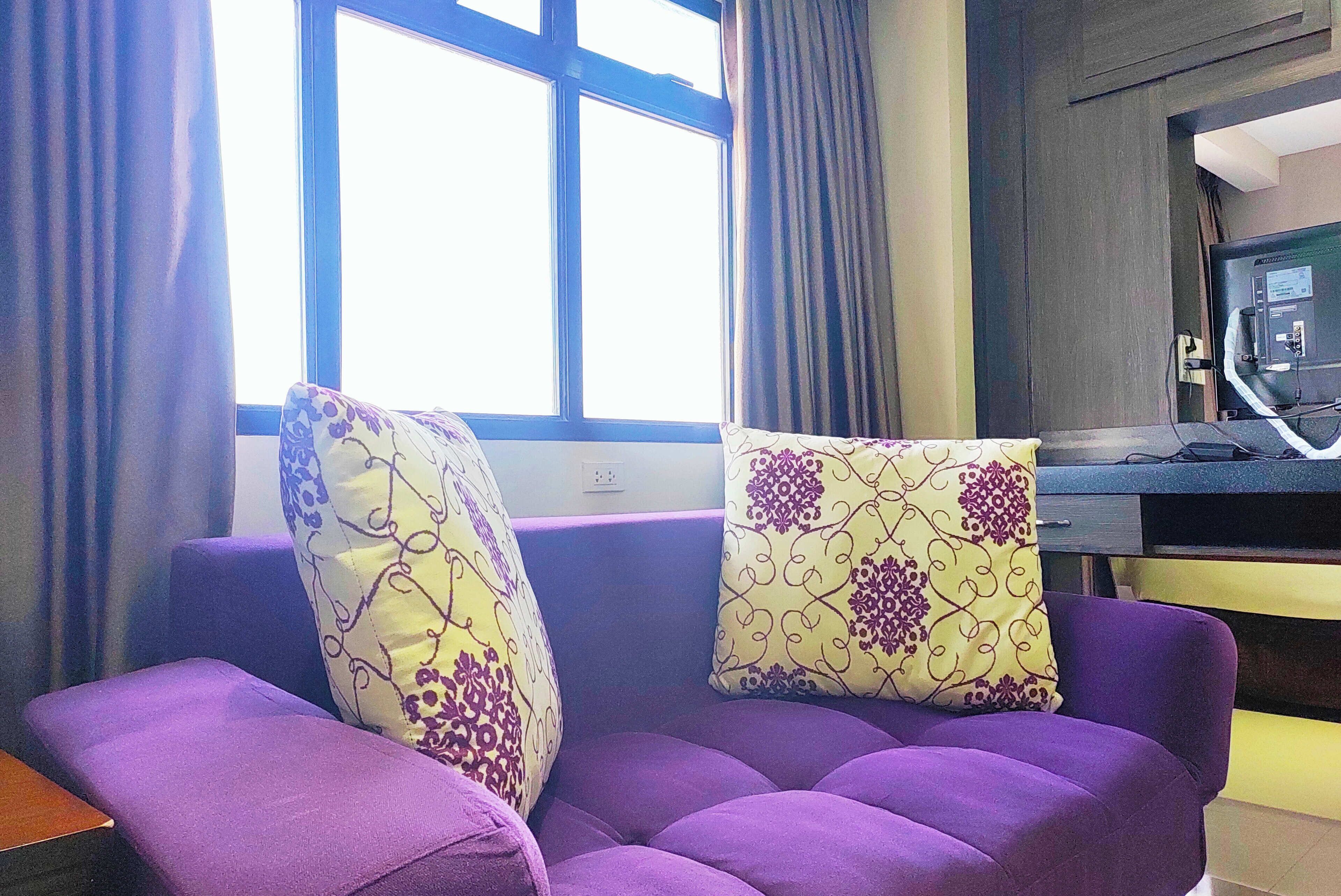 A room with a purple sofa set, two patterned pillows, a desk with a computer, and a window with curtains.