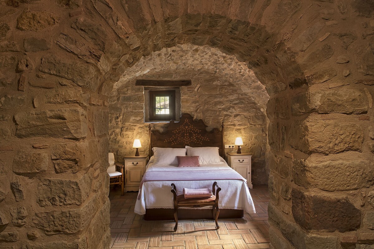 Ein Schlafzimmer mit Steinwänden, einem Bett, zwei Nachttischen, einem Stuhl und einem Fenster.