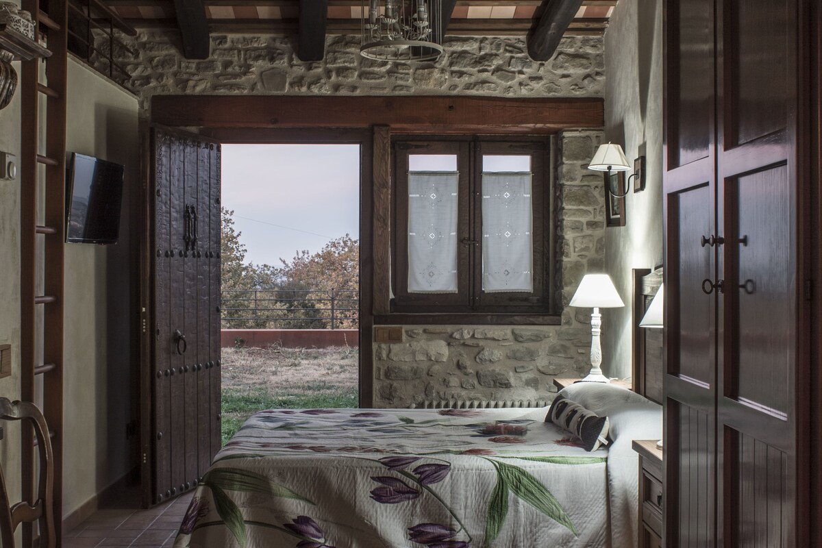 Ein Schlafzimmer mit Steinsandsteinwand, Holzbalken, einem Bett mit floralem Überwurf, einem Nachttisch mit Lampe und einem Fernseher an der Wand.