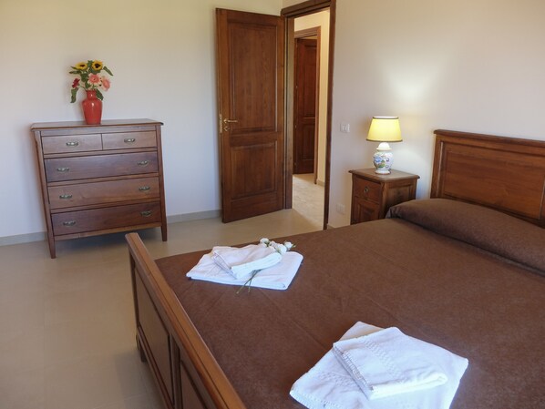 A bedroom with a wooden bed, a dresser, a nightstand with a lamp, and a vase with flowers on the dresser. - Terrazze sul Tirreno (Lascari)