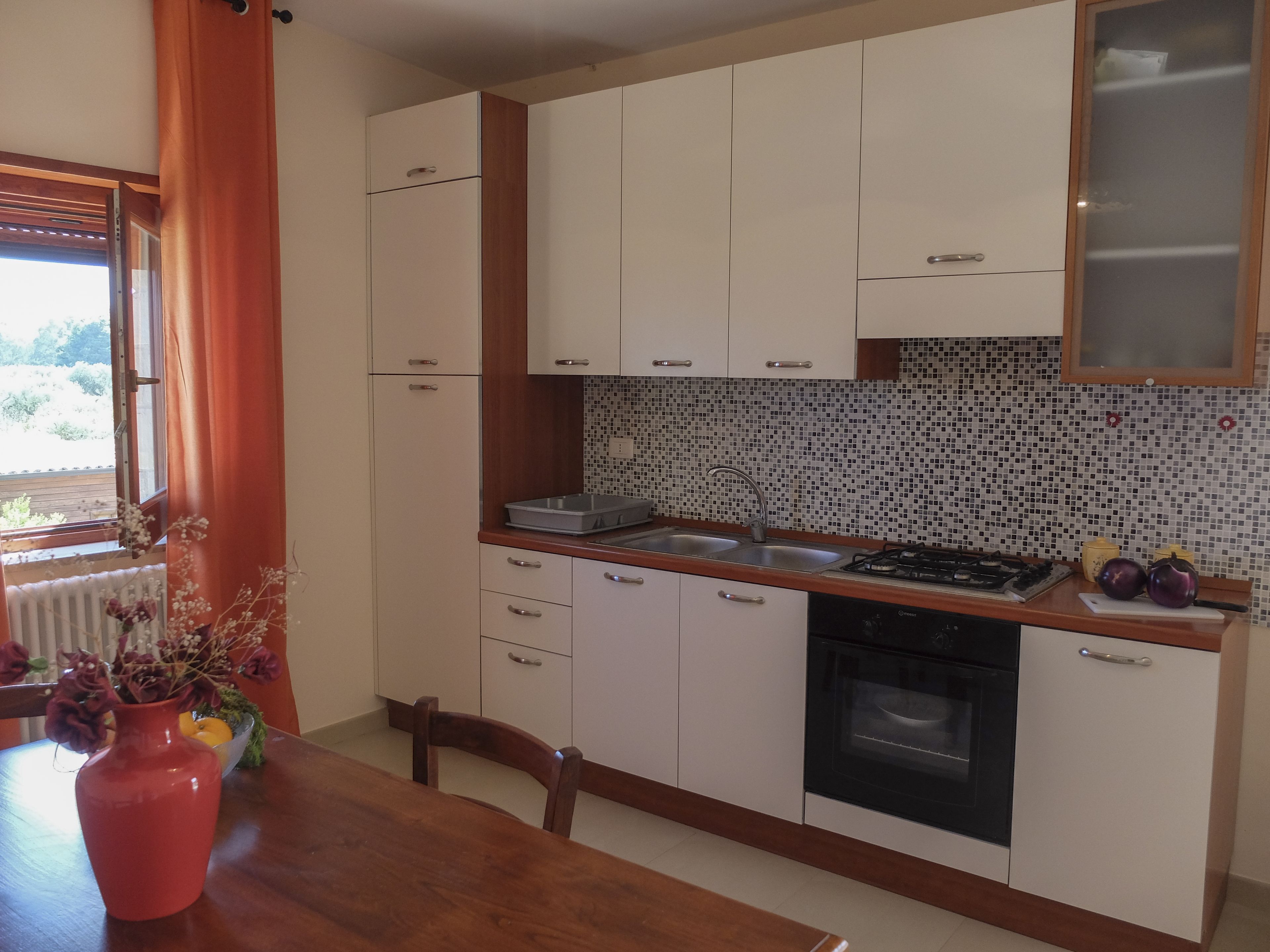 Een keuken met witte kastjes, een betegelde spatwand, een gasfornuis en een raam met gordijnen.