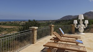 A terrace with wooden lounge chairs and a white candelabra, overlooking a coastal landscape with mountains in the distance. - Terrazze sul Tirreno (Lascari)