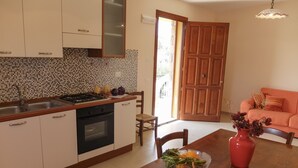 A kitchen with a stove, sink, and cabinets, a dining table with a vase and a bowl of fruit, and a wooden door leading to a balcony. - Terrazze sul Tirreno (Lascari)