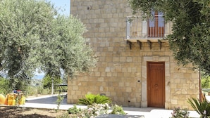 A stone building with a wooden door, a balcony with metal railings, and surrounding greenery including olive trees and flowering plants. - Terrazze sul Tirreno (Lascari)