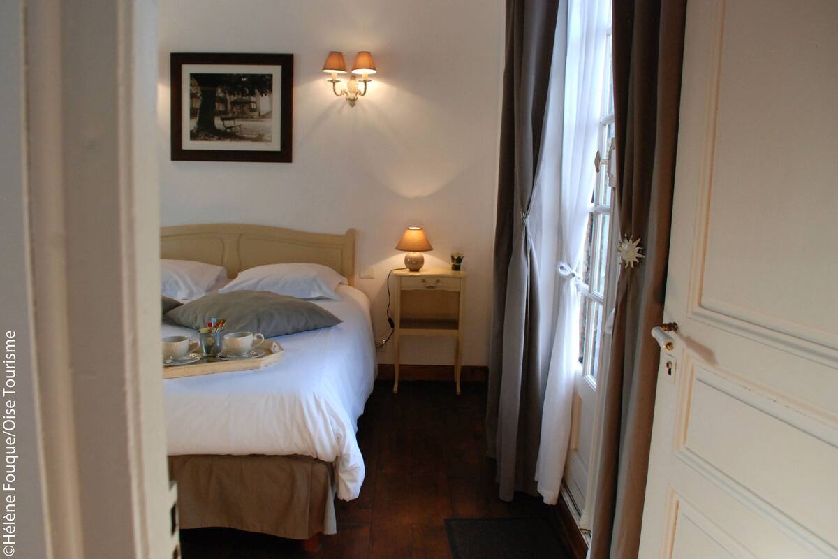A hotel room with a bed, bedside table with a lamp, a framed picture on the wall, and a window with curtains.