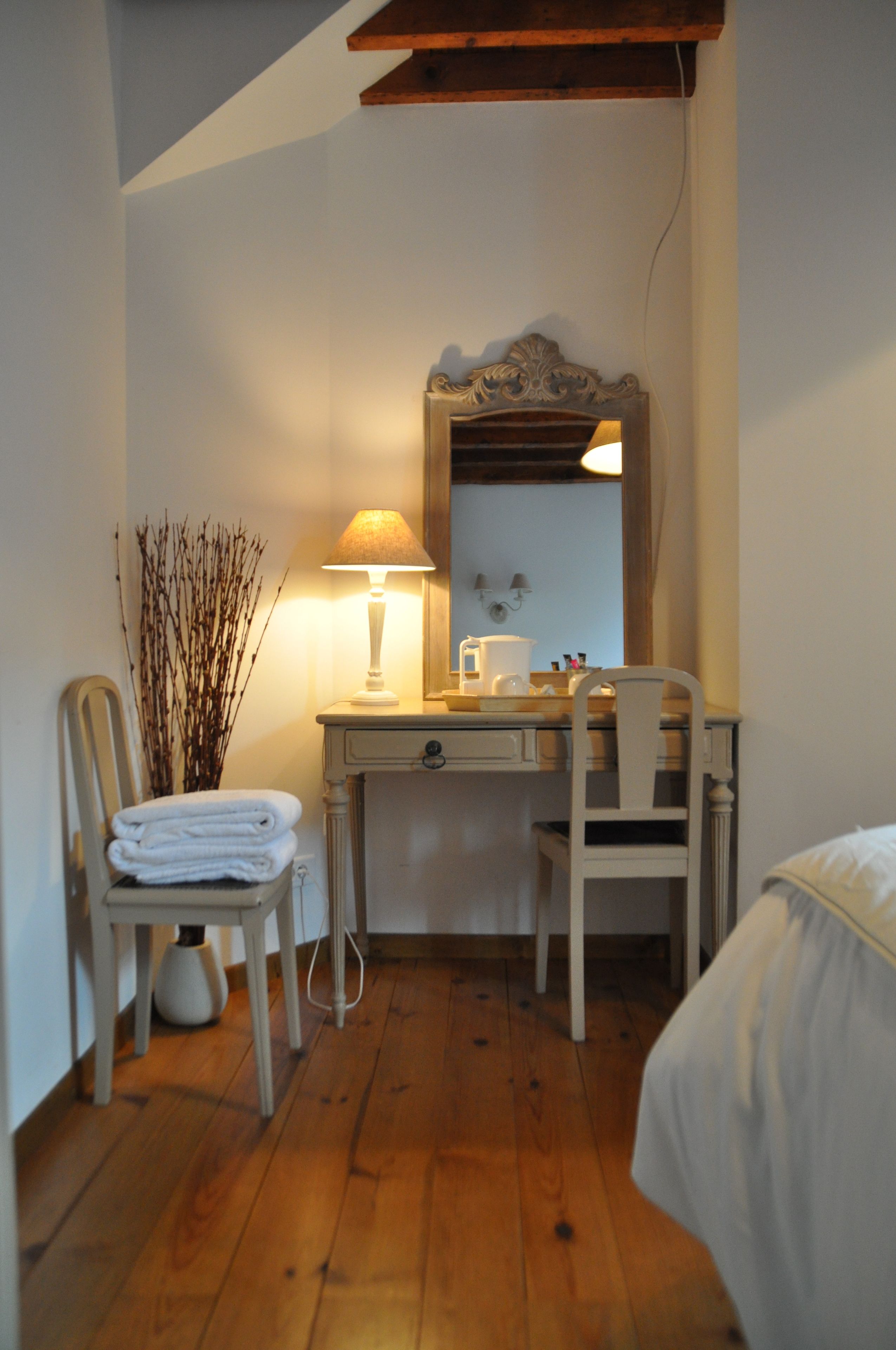 Una habitación pequeña y acogedora con un escritorio de madera, una silla, una lámpara y un espejo. Se ve parcialmente una cama a la derecha.