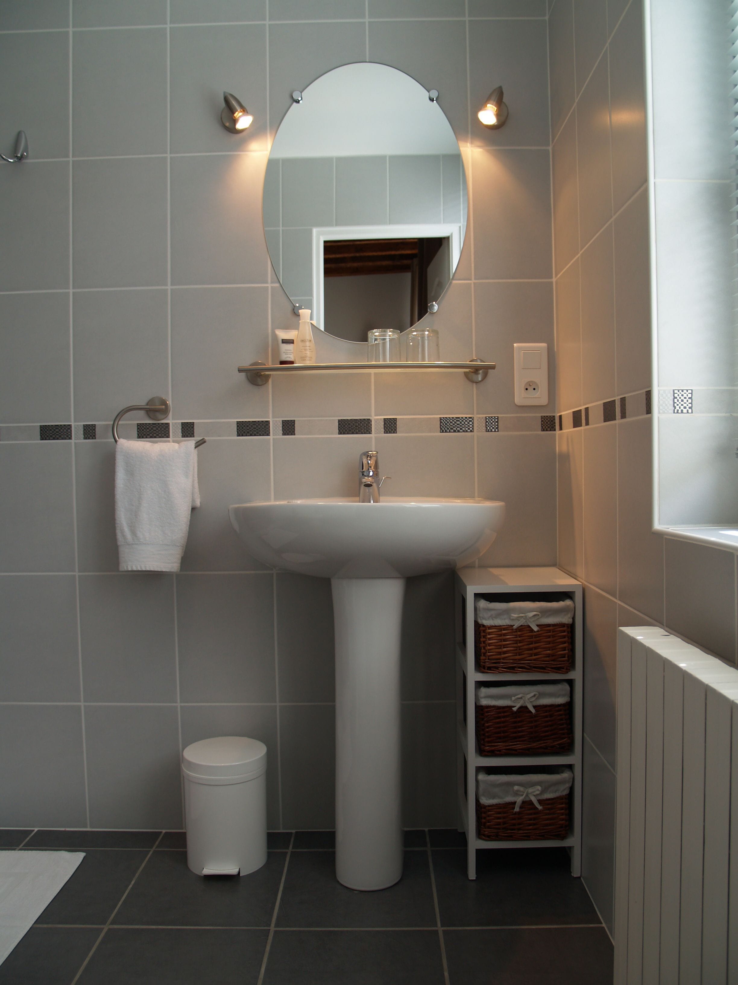 Een badkamer met een wit wastafeltje, een spiegel erboven, een handdoekrek, een prullenbak en rieten manden op een plank.