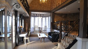 A spacious room with a high ceiling, stone walls, and a large window. There is a staircase with ornate metal railings and a dining area with a table and chairs. - Le Logis De Gerberoy (Gerberoy)