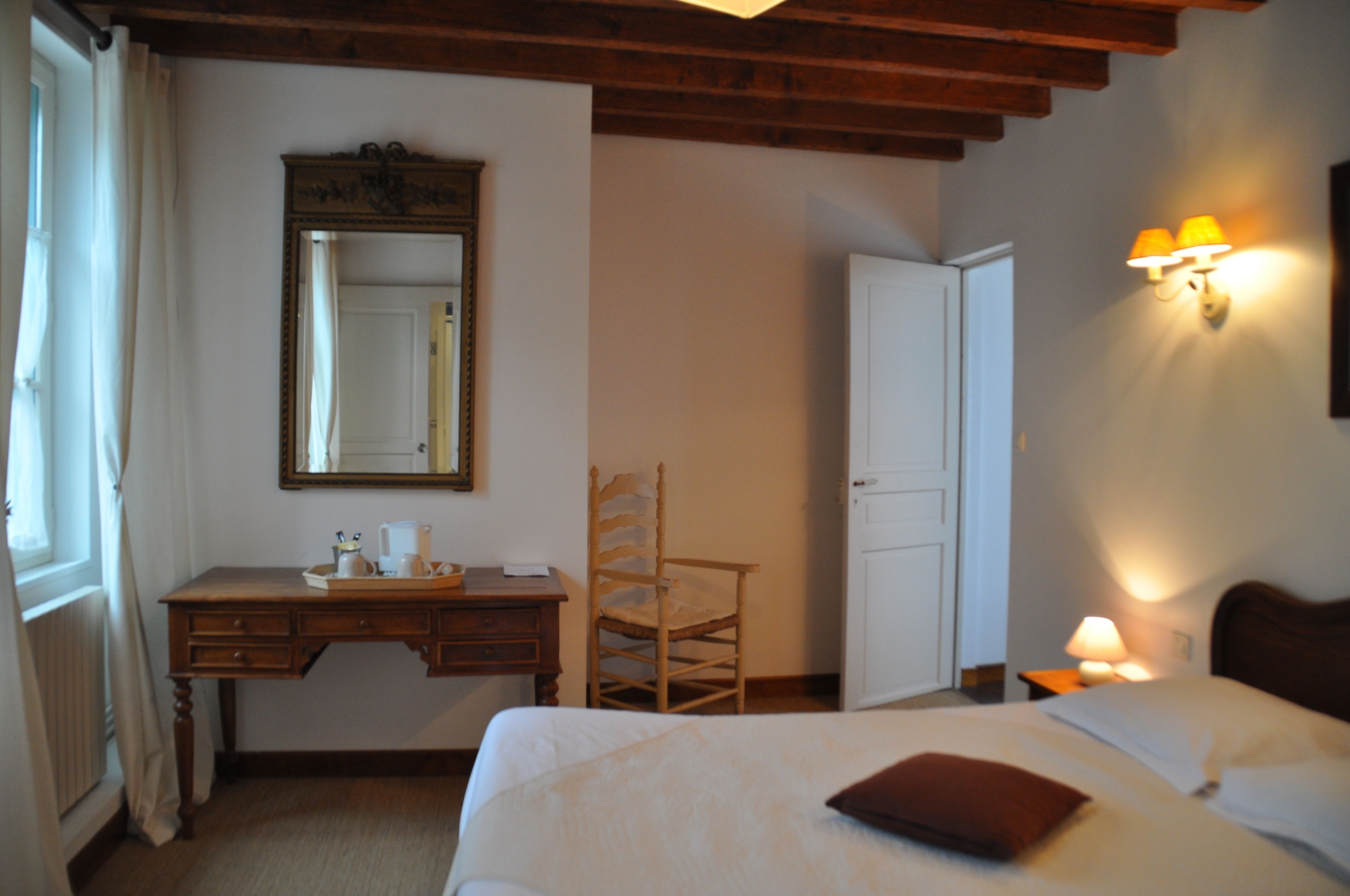 A bedroom with a wooden desk, a chair, a bed with pillows, a nightstand with a lamp, and a wall-mounted mirror.