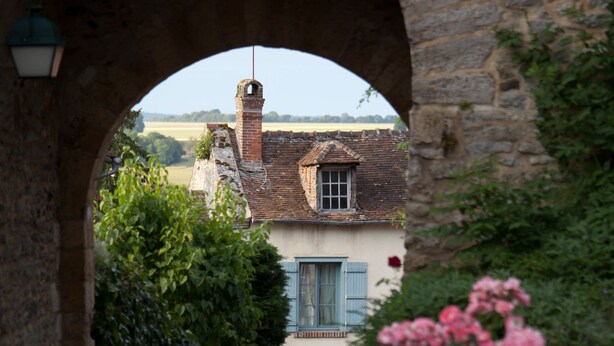 A narrow cobblestone path leading through a stone archway, flanked by lush greenery and a rustic building with a visible window and door. - Le Logis De Gerberoy (Gerberoy)