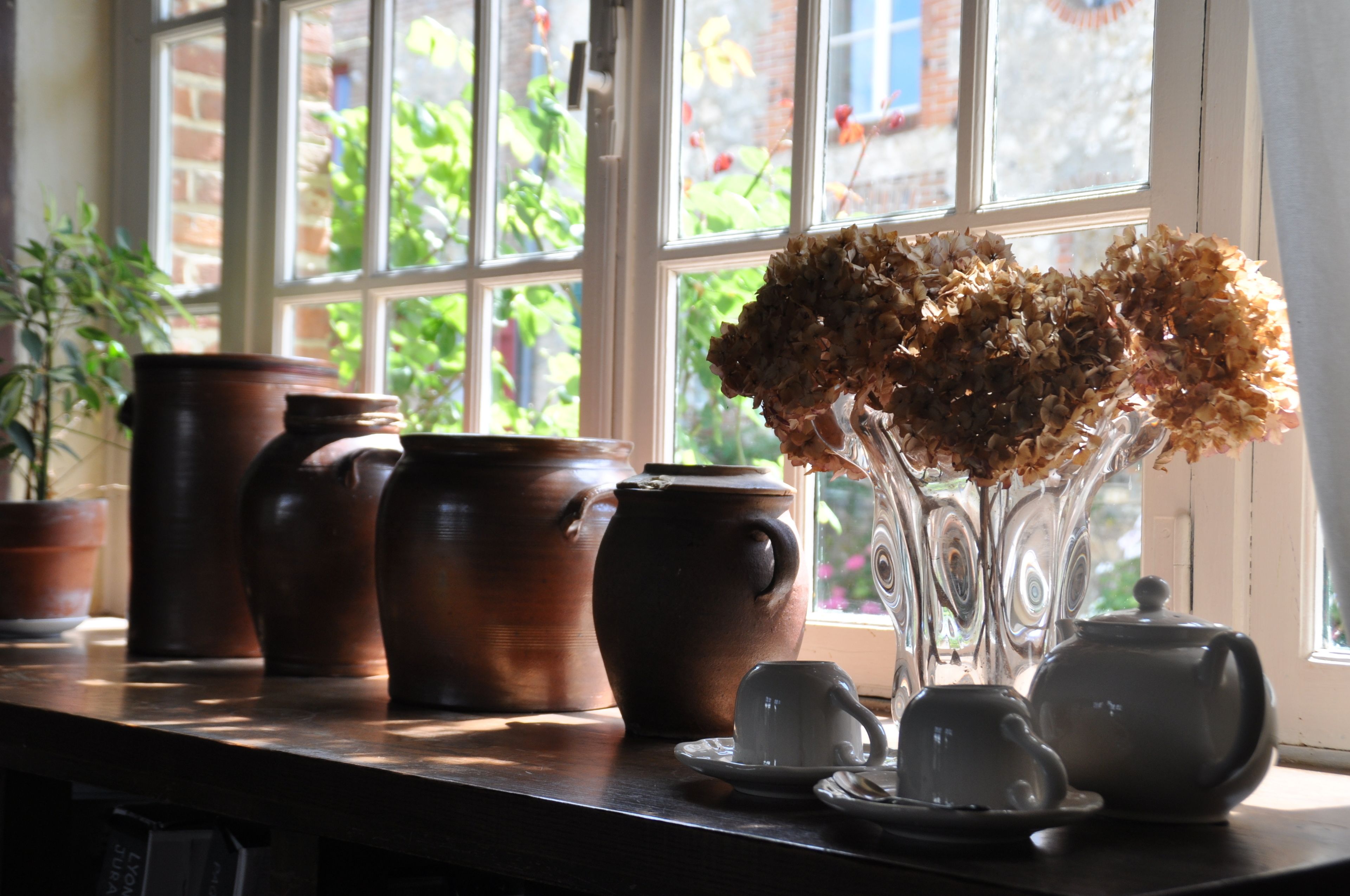 A table with pottery and a vase of dried flowers by a window.