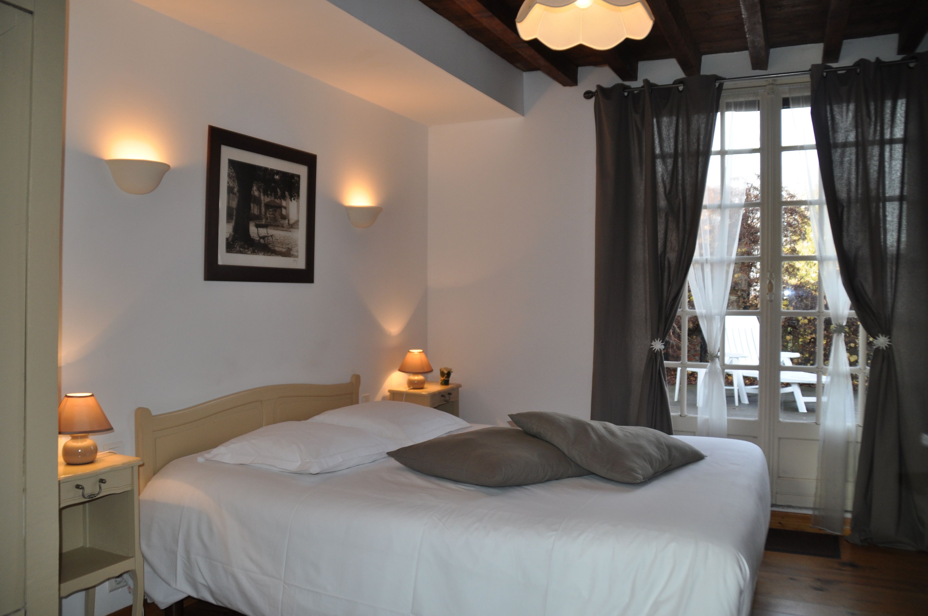 A bedroom with a bed, bedside lamps, a framed picture on the wall, and a view of an outdoor area through a window with curtains.
