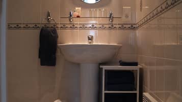 A bathroom with a white sink, a mirror, and a towel rack. There is a trash bin and a shelf with towels.