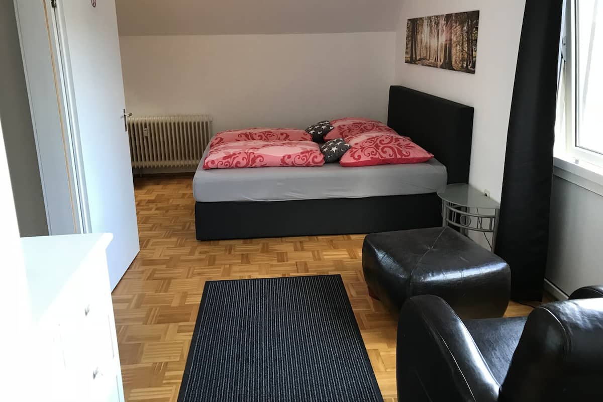 Un dormitorio con una cama, un reposapiés de cuero negro, una mesita pequeña y un radiador.