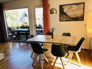 Un coin repas avec une table en bois, des chaises noires et une vue sur un immeuble à travers des portes vitrées.
