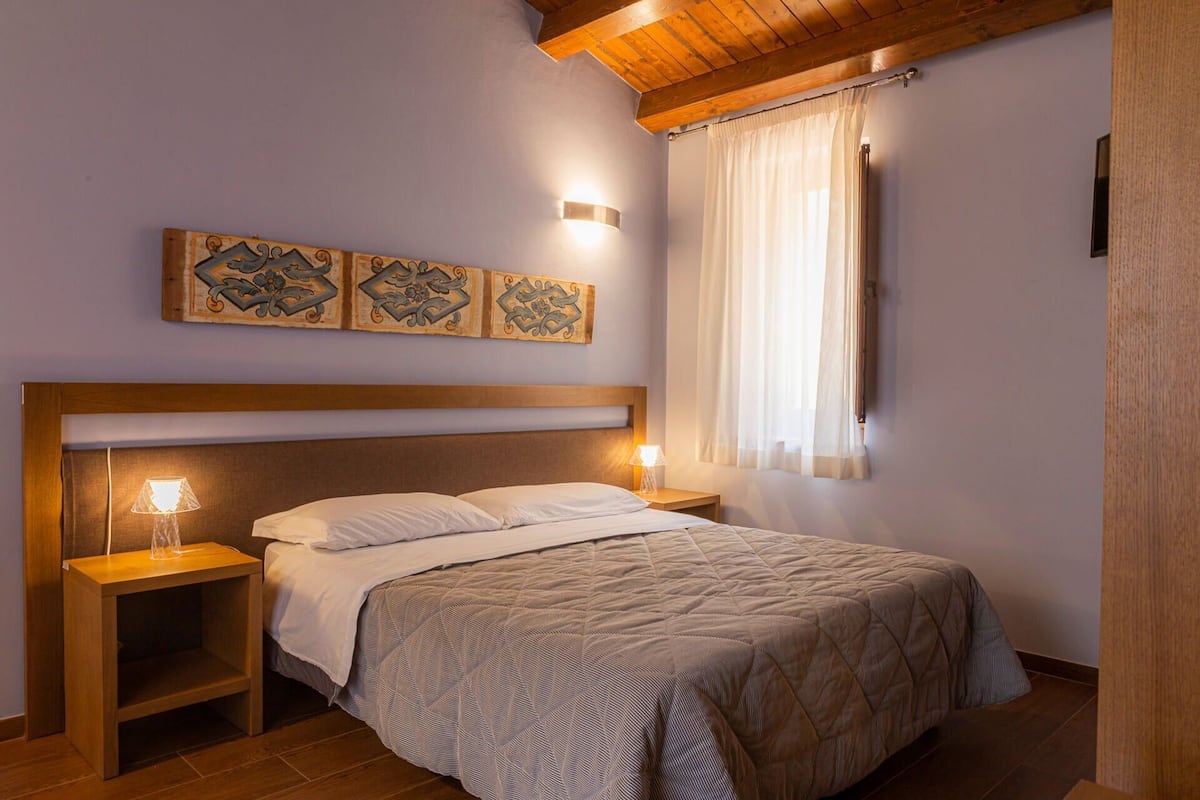 A bedroom with a wooden headboard, a bed with a patterned bedspread, two bedside lamps, and decorative wall art.