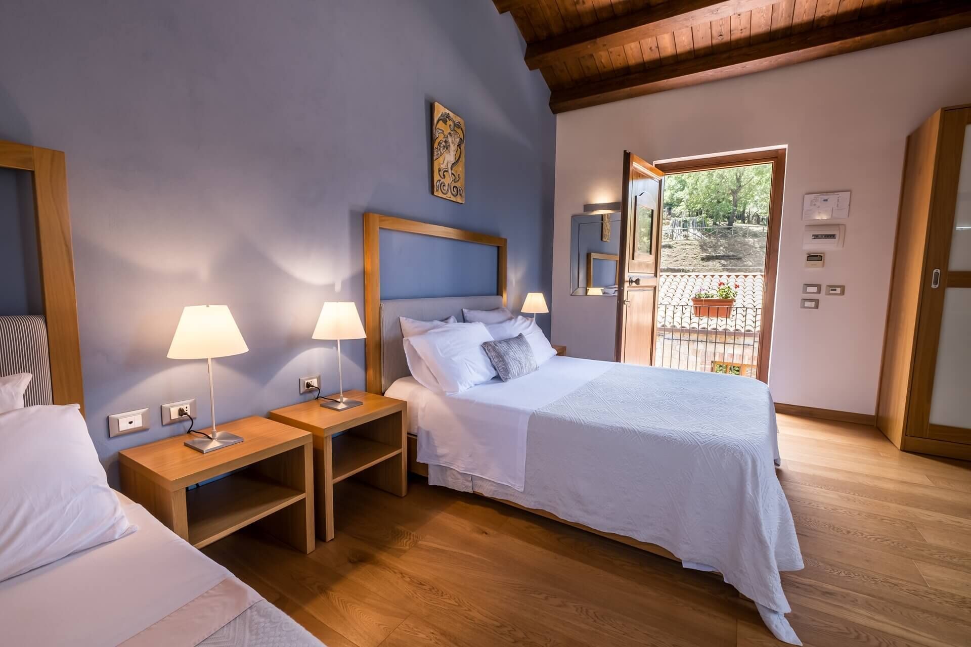 Un dormitorio con una cama, dos mesitas de noche con lámparas, un cabecero de madera y vistas al exterior a través de una puerta.
