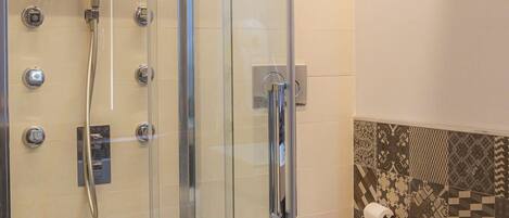 A glass-enclosed shower stall with a handheld showerhead, a toilet, and patterned tiles on the wall.