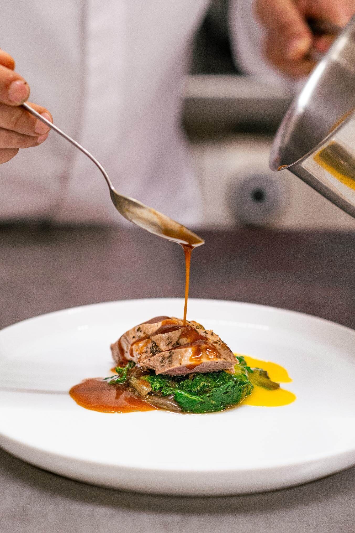 A chef pouring sauce over a dish on a white plate.