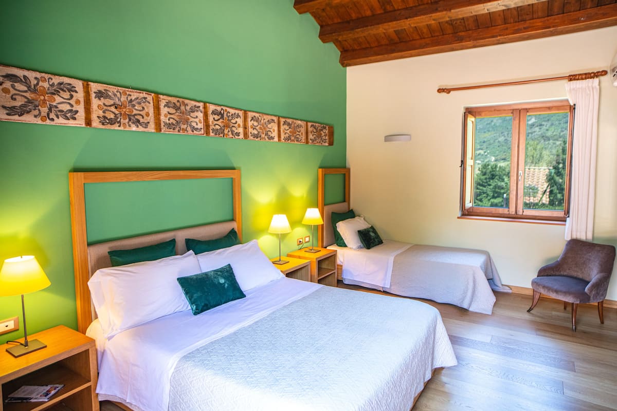 A hotel room with two beds, green walls, wooden headboards, bedside lamps, and a window with a view of greenery.