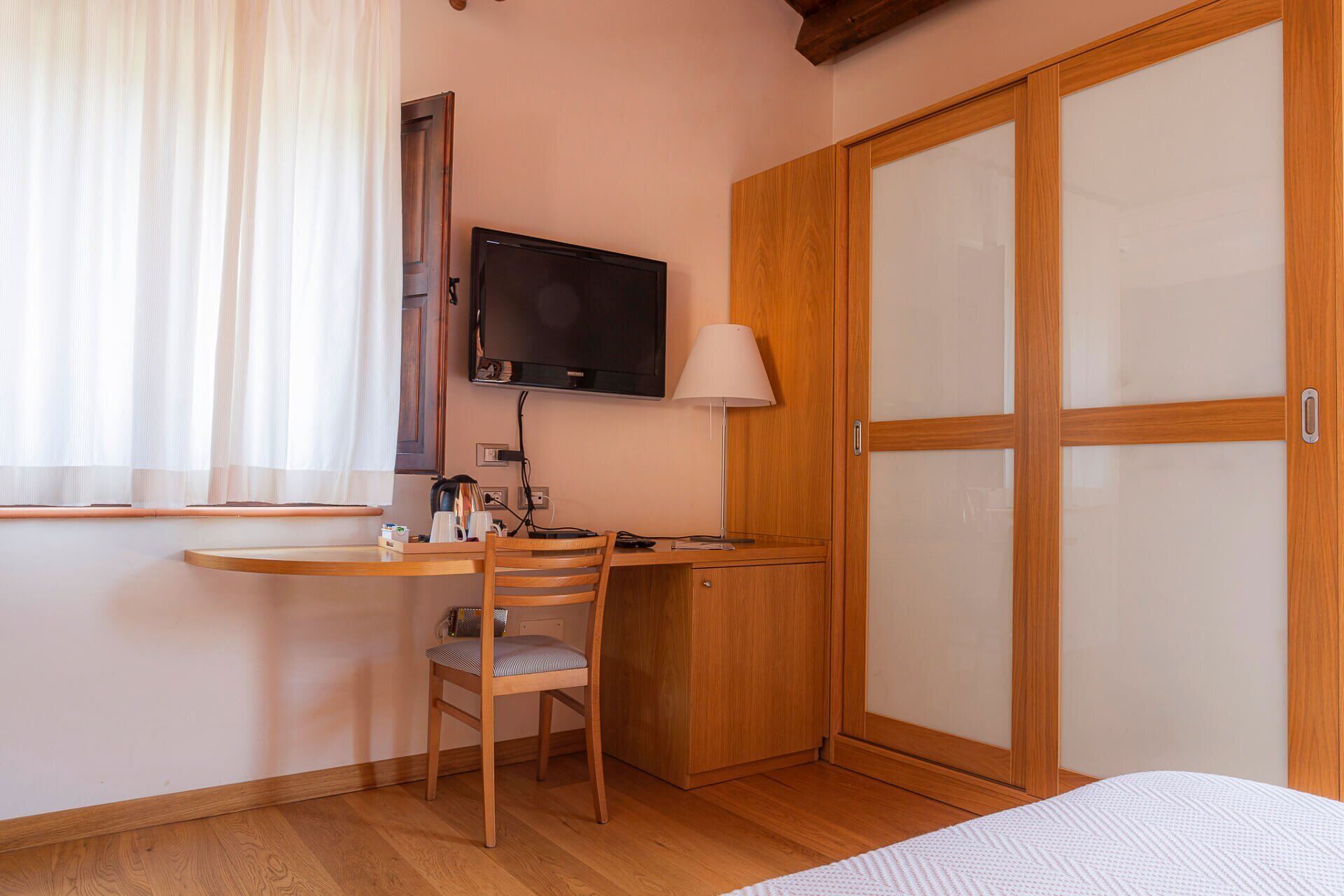 A small room with a wooden desk, a chair, a television mounted on the wall, a lamp, and a wardrobe with a sliding door.