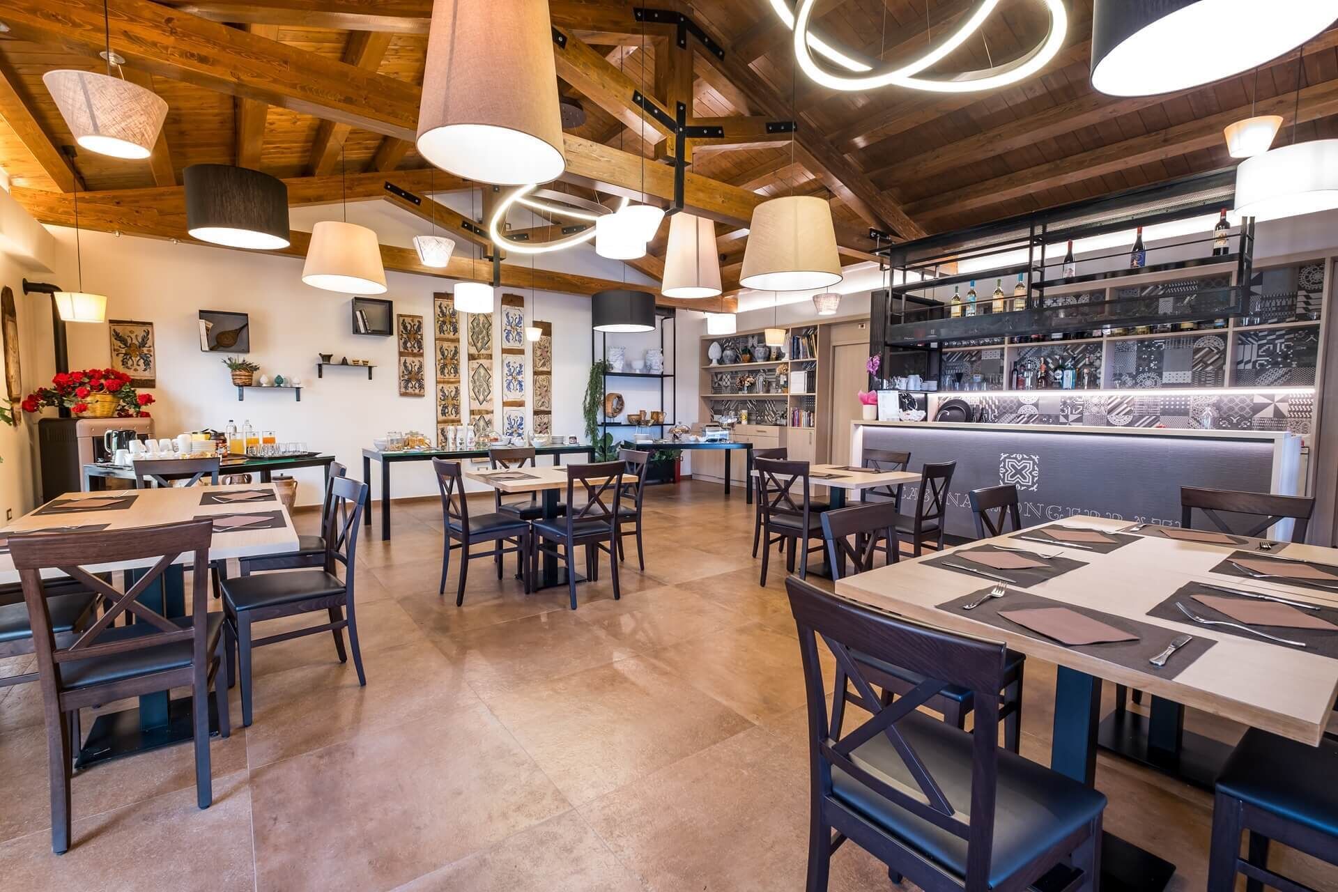 A restaurant with wooden ceiling beams, pendant lighting, and a bar area with shelves of bottles. Tables and chairs are arranged for dining.