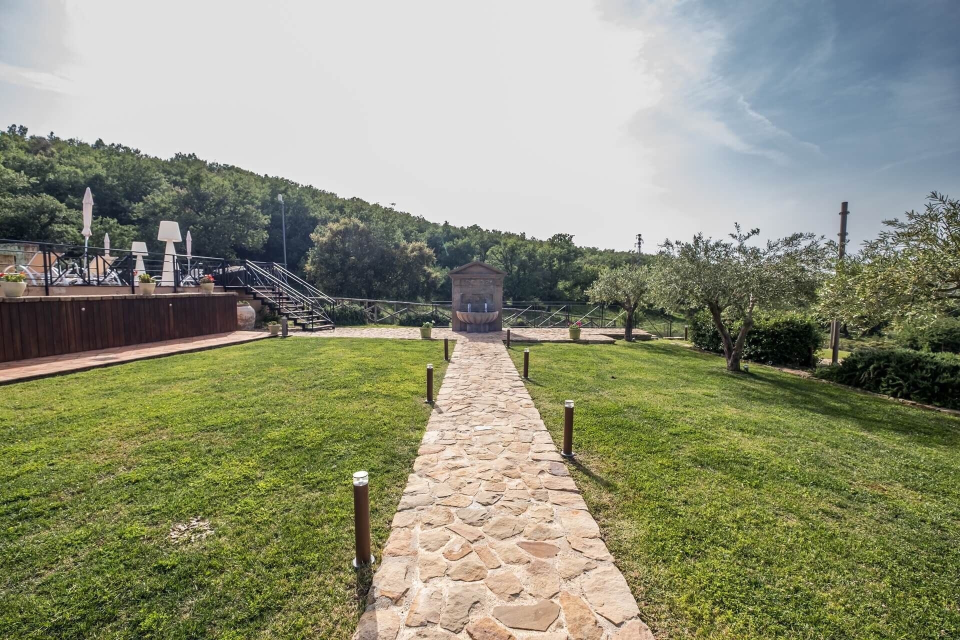 Un sendero de piedra conduce a un pabellón en un parque, con una terraza de madera y asientos al aire libre a la izquierda.