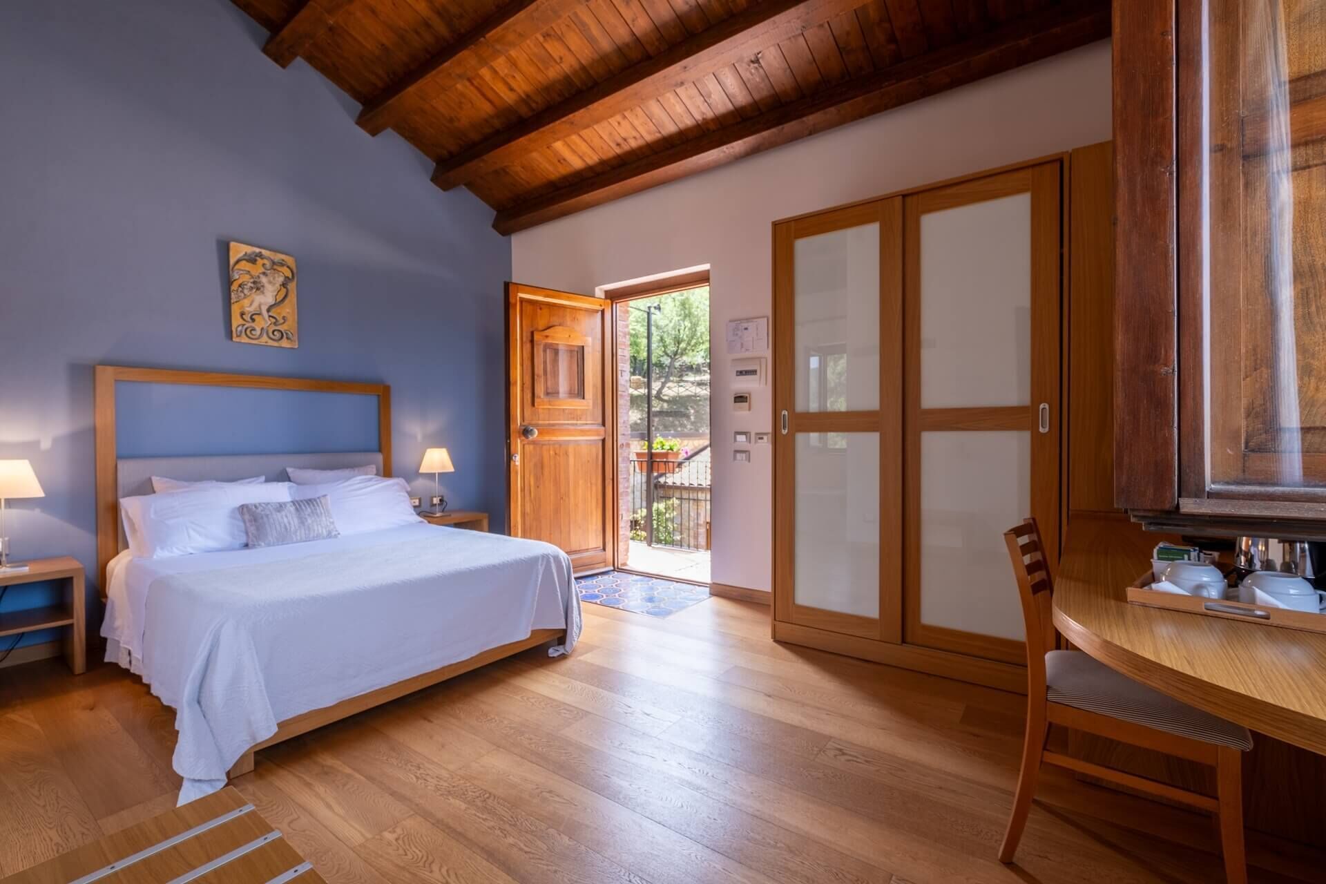 A bedroom with a bed, bedside tables, a wooden ceiling, a door leading outside, and a chair near a table.