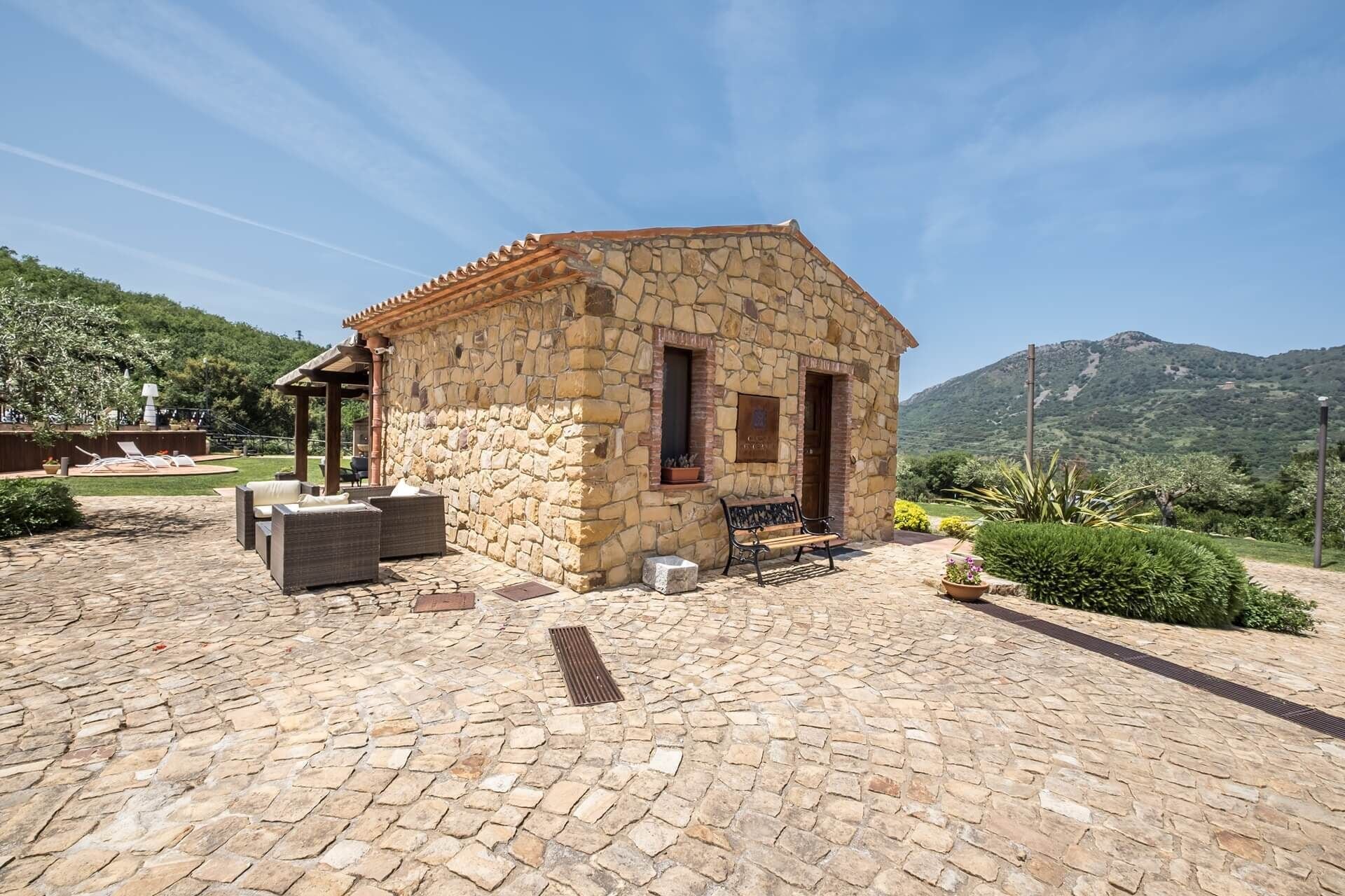A stone building with a tiled roof, a covered patio area with outdoor furniture, and a mountainous backdrop.