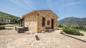 A stone building with a tiled roof, a covered patio area with outdoor furniture, and a mountainous backdrop.